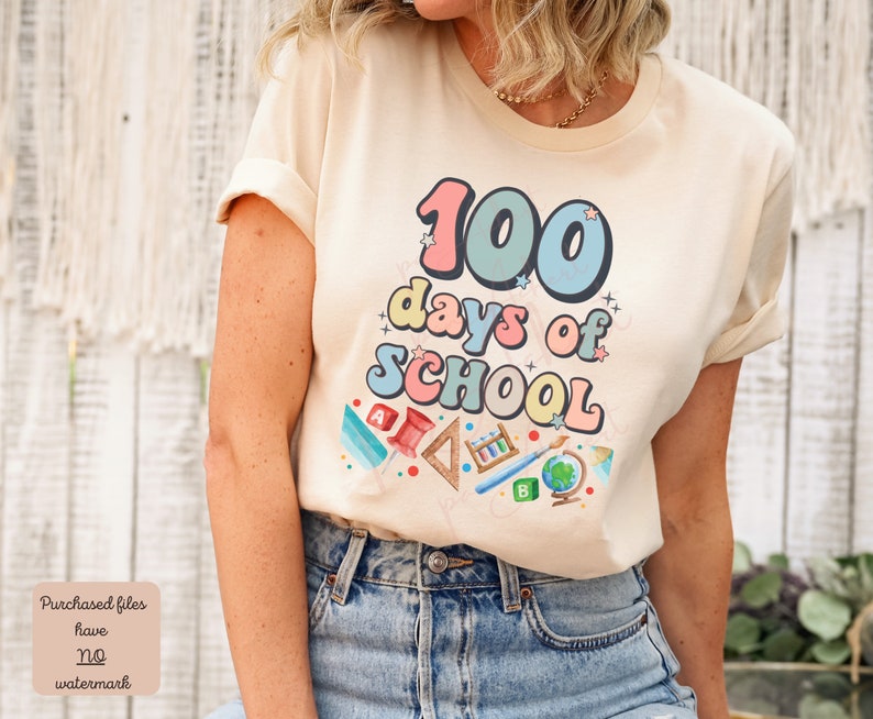 K&ouml;nnte beinhalten: Ein beiges T-Shirt mit einer bunten Grafik, auf der "100 Tage Schule" mit Schulutensilien und Sternen steht.