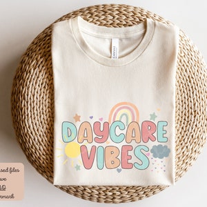 May include: Cream-colored t-shirt with the words "DAYCARE VIBES" in colorful bubble letters. The design includes a rainbow, sun, cloud, and stars. The shirt is displayed on a woven wicker surface.