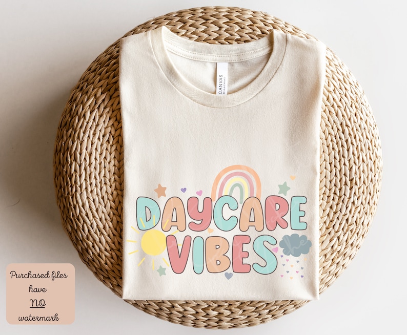 May include: Cream-colored t-shirt with the words "DAYCARE VIBES" in colorful bubble letters. The design includes a rainbow, sun, cloud, and stars. The shirt is displayed on a woven wicker surface.