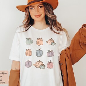 May include: A white t-shirt with a pattern of pastel colored pumpkins. The pumpkins are in various shades of pink, blue, and green. The shirt is a simple design and would be perfect for fall.