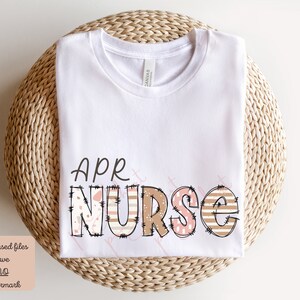May include: White t-shirt featuring the text "APR NURSE" in brown, pink and white patterned letters. The letters have a stitched appearance.