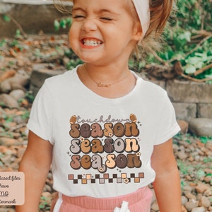 May include: A young girl wearing a white t-shirt with a brown, orange, and yellow graphic that says "touchdown season season season". The graphic also includes a football and flowers.