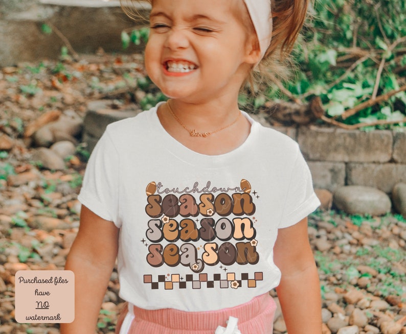 May include: A young girl wearing a white t-shirt with a brown, orange, and yellow graphic that says "touchdown season season season". The graphic also includes a football and flowers.