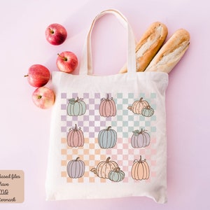 May include: A white canvas tote bag with a checkered pattern featuring pastel-coloured pumpkins. The bag has a long handle and is filled with two baguettes and apples.
