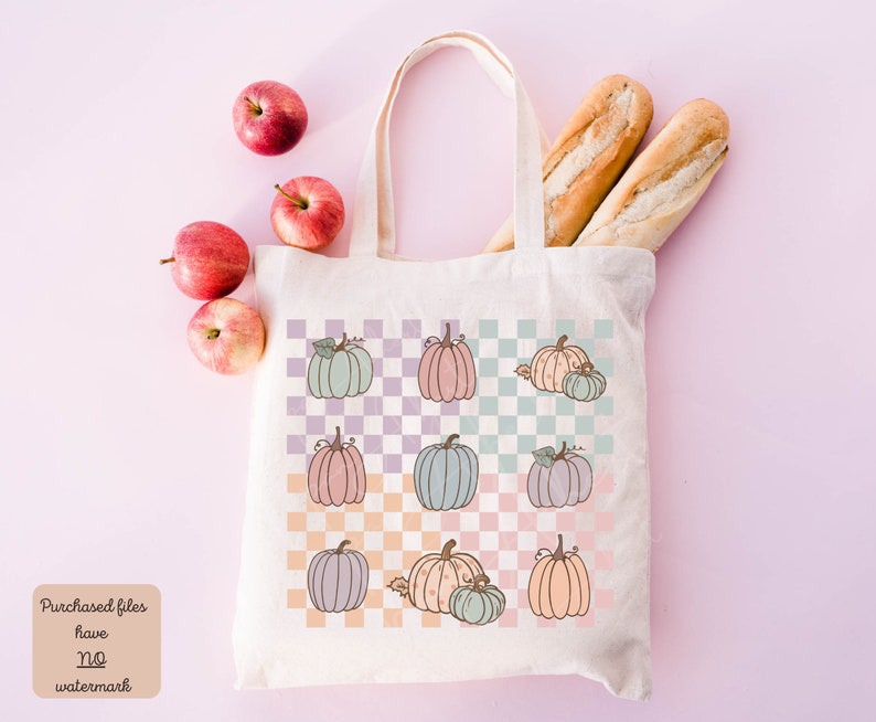 May include: A white canvas tote bag with a checkered pattern featuring pastel-coloured pumpkins. The bag has a long handle and is filled with two baguettes and apples.