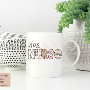May include: White ceramic mug with a brown and pink graphic that says "APR NURSE". The mug has a black and white striped handle.
