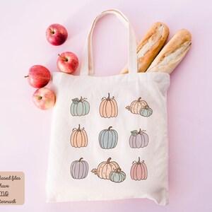 May include: A white canvas tote bag with a design of nine pumpkins in various pastel colors. The pumpkins are arranged in three rows of three. The bag is sitting on a pink surface with apples and baguettes.