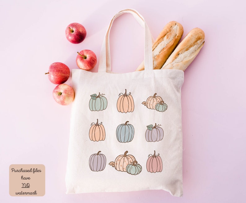 May include: A white canvas tote bag with a design of nine pumpkins in various pastel colors. The pumpkins are arranged in three rows of three. The bag is sitting on a pink surface with apples and baguettes.