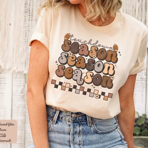 May include: A beige t-shirt with a retro-style graphic that says "Touchdown Season" in brown, gray, and beige letters. The graphic also includes footballs and flowers.