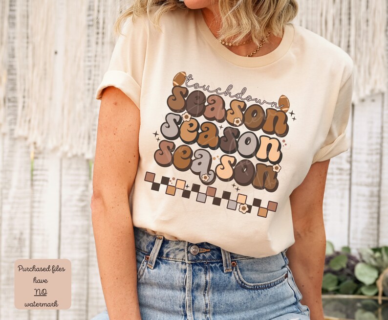 May include: A beige t-shirt with a retro-style graphic that says "Touchdown Season" in brown, gray, and beige letters. The graphic also includes footballs and flowers.