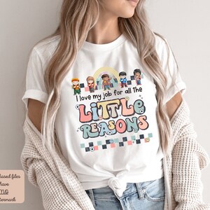 Pu&ograve; includere: Maglietta bianca con una grafica colorata che dice "I love my job for all the little reasons". La grafica presenta un arcobaleno, un gruppo di bambini e uno schema a scacchi.