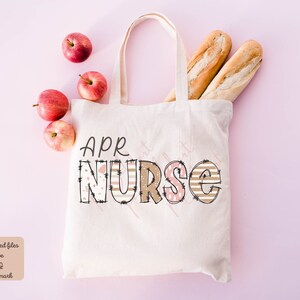 May include: A white canvas tote bag featuring the text "APR NURSE" in brown and pink patterned letters. The bag is displayed against a pink background with apples and bread.
