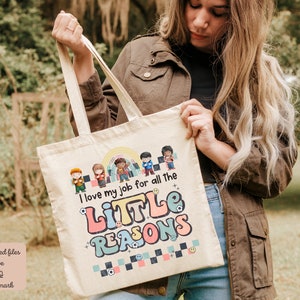 Pu&ograve; includere: Una borsa tote in tela bianca con un design grafico colorato che dice "Adoro il mio lavoro per tutte le piccole ragioni". La grafica include una fila di bambini dei cartoni animati con diversi toni della pelle e acconciature.