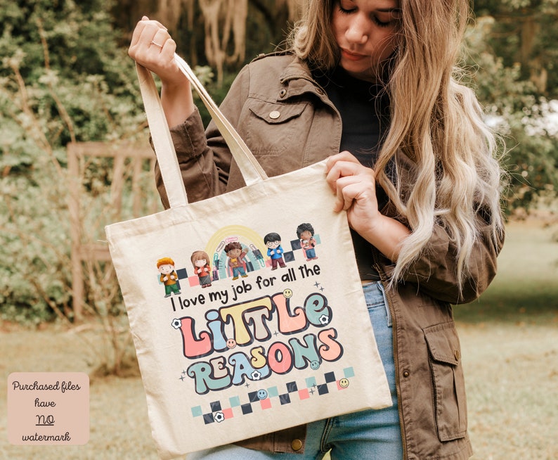 Pu&ograve; includere: Una borsa tote in tela bianca con un design grafico colorato che dice "Adoro il mio lavoro per tutte le piccole ragioni". La grafica include una fila di bambini dei cartoni animati con diversi toni della pelle e acconciature.