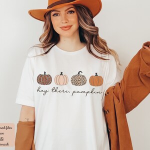 May include: White t-shirt with a graphic of four pumpkins. The pumpkins are in a row and have different patterns, including leopard print and solid colors. The text "hey there, pumpkin" is written below the pumpkins.