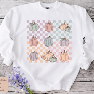 May include: White sweatshirt featuring a checkered pattern in pastel colours. The pattern includes multiple illustrations of pumpkins in various shades of pink, green, and orange.