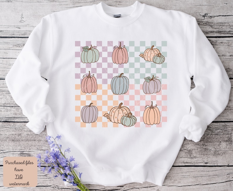 May include: White sweatshirt featuring a checkered pattern in pastel colours. The pattern includes multiple illustrations of pumpkins in various shades of pink, green, and orange.