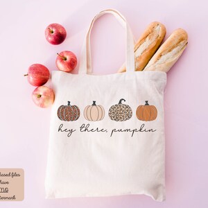 May include: A white canvas tote bag with the text "Hey there, pumpkin" printed on it. The bag features four pumpkins with different patterns, including leopard print and solid colors.