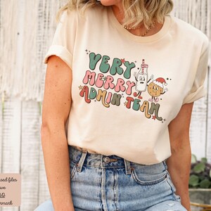 May include: A cream colored t-shirt with a retro design featuring a milk carton and a cookie wearing a Santa hat. The text on the shirt reads "Very Merry Admin Team".