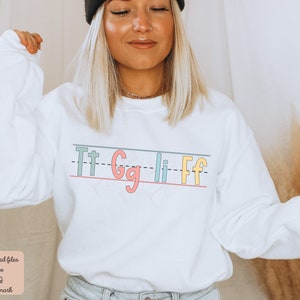 Pu&ograve; includere: Felpa bianca con una grafica delle lettere "Tt Gg Li Ff" scritte in uno stile calligrafico colorato su uno sfondo rigato.