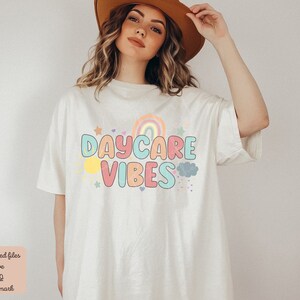 May include: Cream-colored t-shirt with the words "DAYCARE VIBES" in colorful bubble letters. The design includes a rainbow, sun, stars, and clouds. The shirt is a casual, oversized fit.