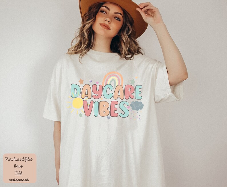 May include: Cream-colored t-shirt with the words "DAYCARE VIBES" in colorful bubble letters. The design includes a rainbow, sun, stars, and clouds. The shirt is a casual, oversized fit.