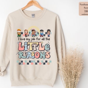 Pu&ograve; includere: Una felpa beige con un design grafico che presenta un gruppo di bambini e il testo "Amo il mio lavoro per tutte le PICCOLE RAGIONI".