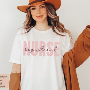 Puede incluir: Una camiseta blanca con el texto "NURSE registered" en rosa con una textura brillante.