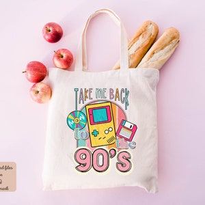 K&ouml;nnte beinhalten: Eine wei&szlig;e Canvas-Tragetasche mit dem Text "Take Me Back to the 90's" in bunter Retro-Schrift. Die Tasche zeigt eine Grafik einer alten Videospielkonsole, einer Diskette und einer CD.