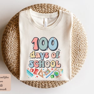 K&ouml;nnte beinhalten: Ein beiges T-Shirt mit dem Text "100 Tage Schule" in bunten, Retro-Lettern. Das Shirt zeigt verschiedene schulthemenbezogene Illustrationen, darunter einen Globus, Bleistifte und ein Lineal.
