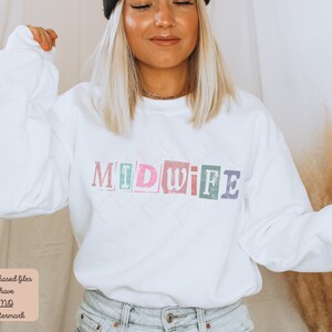 May include: White sweatshirt with the word "MIDWIFE" printed in colorful, distressed letters. The letters are arranged in a staggered pattern.