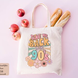 May include: A white canvas tote bag with the text "Take Me Back to the 90's" in colorful retro font. The text is surrounded by a colorful sunburst design and flowers.