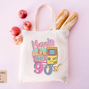 May include: A white canvas tote bag with a retro design featuring the text "Made in the 90's" in pink, yellow, and blue. A yellow and green pixelated video game console is featured on the bag.