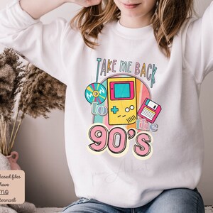K&ouml;nnte beinhalten: Wei&szlig;er Pullover mit einem Grafikdesign, das eine Retro-Videospielkonsole, eine Diskette und eine CD zeigt. Der Text "Take Me Back to the 90's" ist in einer bunten Schrift gedruckt.