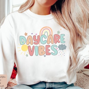 May include: White sweatshirt with the text "DAYCARE VIBES" in colorful bubble letters. The design includes a rainbow, sun, stars, and a cloud with hearts. A fun and cheerful design for daycare staff or anyone who loves working with children.