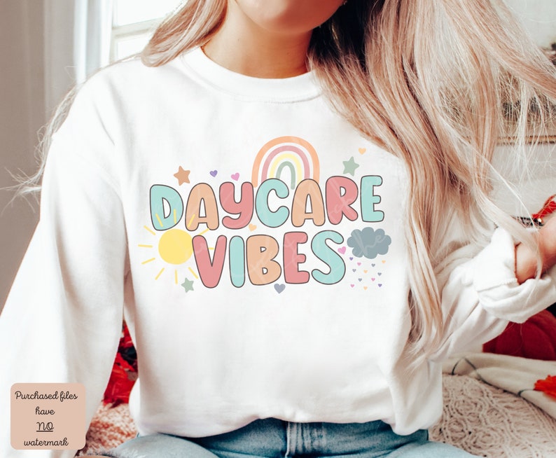 May include: White sweatshirt with the text "DAYCARE VIBES" in colorful bubble letters. The design includes a rainbow, sun, stars, and a cloud with hearts. A fun and cheerful design for daycare staff or anyone who loves working with children.