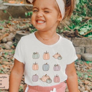 May include: A young girl wearing a white t-shirt with a pattern of pastel colored pumpkins. The shirt has short sleeves and a crew neck. The girl is smiling and has a headband on.