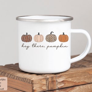 May include: White enamel mug with a handle. The mug has a design of four pumpkins in a row. The pumpkins are orange, brown, and white with a leopard print pattern. The text "Hey there, pumpkin" is written below the pumpkins.