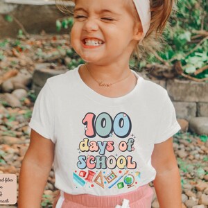 K&ouml;nnte beinhalten: Ein kleines Kind tr&auml;gt ein wei&szlig;es T-Shirt mit dem Text "100 Tage Schule" in bunten Buchstaben. Das Shirt zeigt au&szlig;erdem verschiedene bunte Schulutensilien, darunter einen Bleistift, ein Lineal, einen Globus und ein Buch. Das Kind l&auml;chelt und schaut in die Kamera.