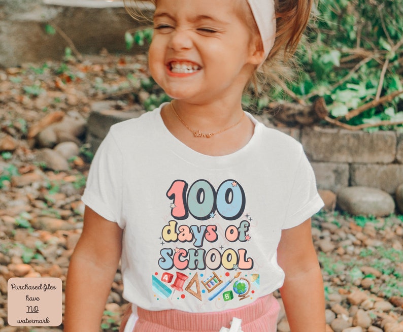 K&ouml;nnte beinhalten: Ein kleines Kind tr&auml;gt ein wei&szlig;es T-Shirt mit dem Text "100 Tage Schule" in bunten Buchstaben. Das Shirt zeigt au&szlig;erdem verschiedene bunte Schulutensilien, darunter einen Bleistift, ein Lineal, einen Globus und ein Buch. Das Kind l&auml;chelt und schaut in die Kamera.