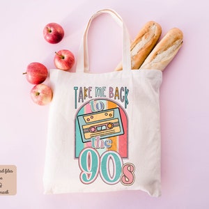 May include: A white tote bag with the text "Take Me Back to the 90s" and a retro cassette tape graphic. The bag is on a pink background with apples and baguettes.