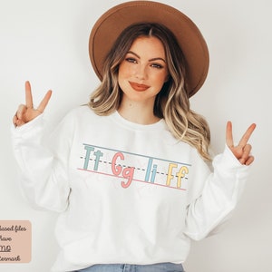 Pu&ograve; includere: Felpa bianca con una grafica delle lettere "Tt Gg Ii Ff" scritte in un font colorato e giocoso, come se fossero su un foglio di carta rigato. Le lettere sono di diversi colori, tra cui rosa, blu, giallo e verde.