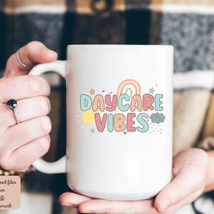 May include: White ceramic mug with the words "DAYCARE VIBES" in colorful, playful lettering. The design includes a rainbow, sun, stars, and clouds. The mug is held by a person.