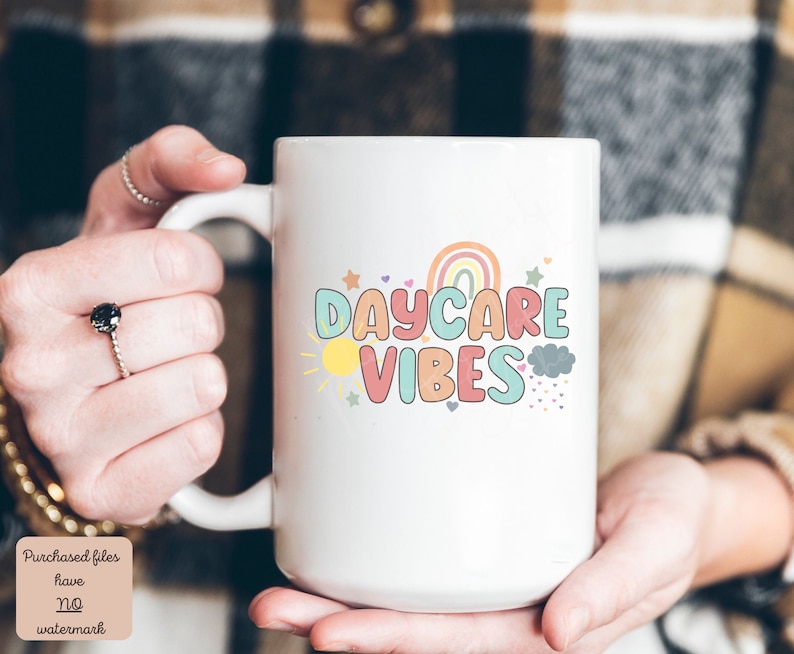 May include: White ceramic mug with the words "DAYCARE VIBES" in colorful, playful lettering. The design includes a rainbow, sun, stars, and clouds. The mug is held by a person.