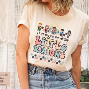 Pu&ograve; includere: Una maglietta beige con una grafica colorata che dice "I love my job for all the little reasons". La grafica presenta un arcobaleno, un pallone da calcio e bambini dei cartoni animati.