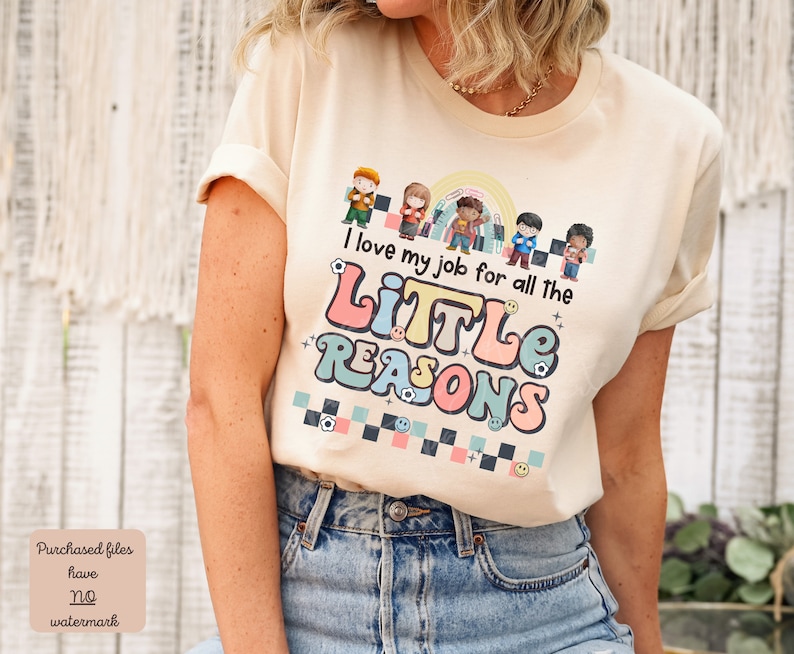 Pu&ograve; includere: Una maglietta beige con una grafica colorata che dice "I love my job for all the little reasons". La grafica presenta un arcobaleno, un pallone da calcio e bambini dei cartoni animati.