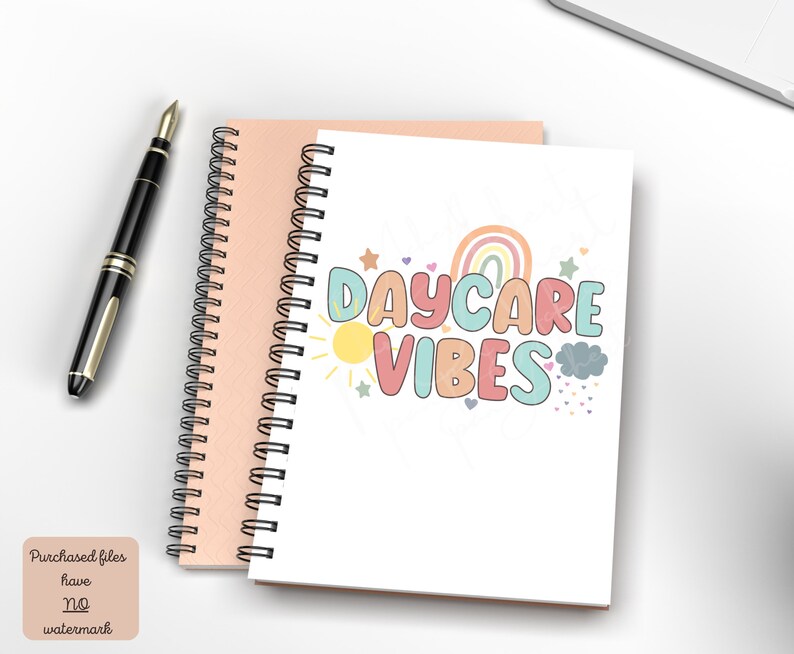 May include: Two spiral-bound notebooks, one peach and one white, with a black pen. The white notebook features the text "DAYCARE VIBES" in colorful bubble letters, with a rainbow, sun, and stars graphic. The notebooks are on a white surface.