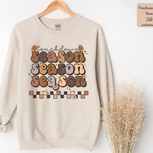 May include: A beige sweatshirt with a graphic design that says "touchdown season" in a retro font with brown, tan, and orange colors. The graphic also includes footballs and flowers.
