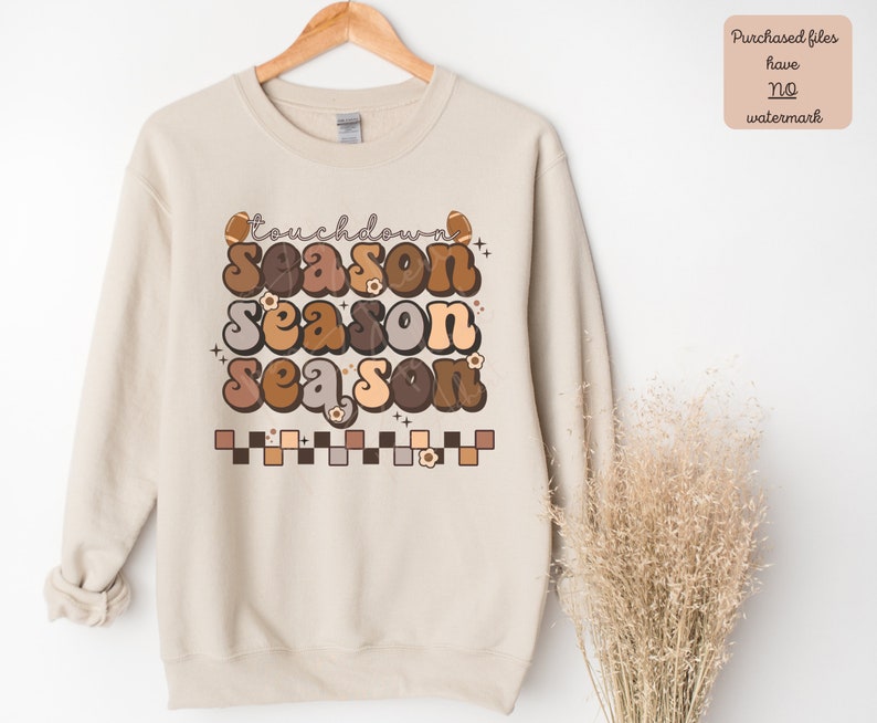 May include: A beige sweatshirt with a graphic design that says "touchdown season" in a retro font with brown, tan, and orange colors. The graphic also includes footballs and flowers.