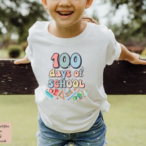 K&ouml;nnte beinhalten: Ein wei&szlig;es T-Shirt mit einer bunten Grafik, auf der "100 Tage Schule" steht, umgeben von Schulutensilien. Das Shirt wird von einem Kind getragen.